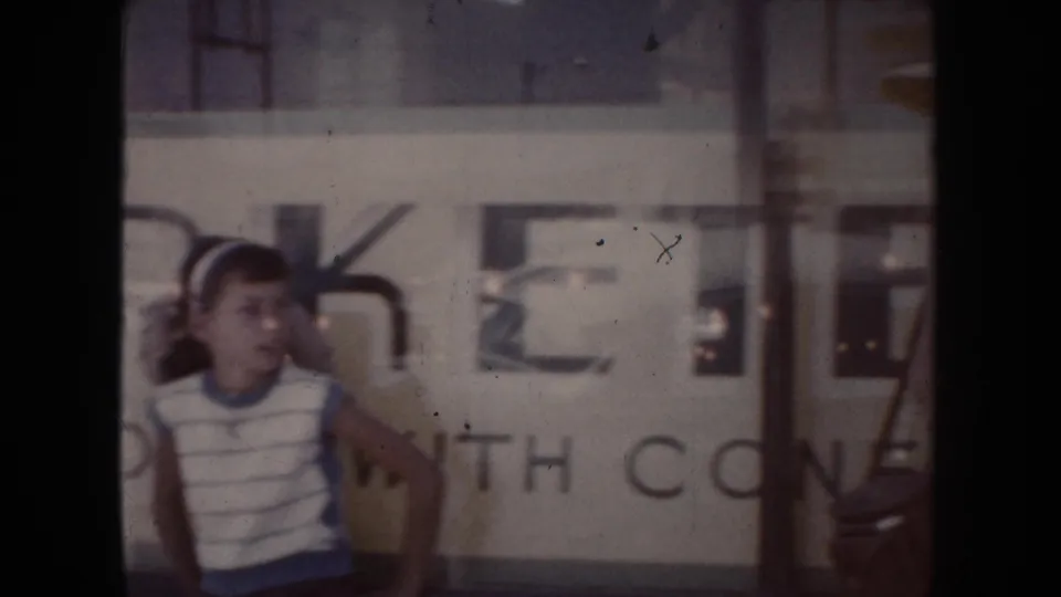 South Dakota home-movie frame from the clip showing a young person in a striped sleeveless shirt near a storefront with partial lettering.