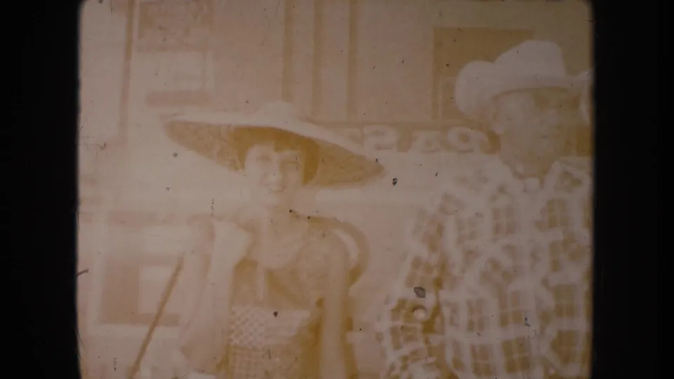 Faded 1954 South Dakota frame showing the woman in a straw hat and the man in a cowboy hat standing together near storefront signage.