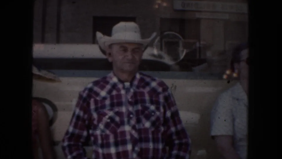 In 1954 South Dakota footage, a man in a white cowboy hat and plaid shirt stands beside a parked car with another person partly visible.