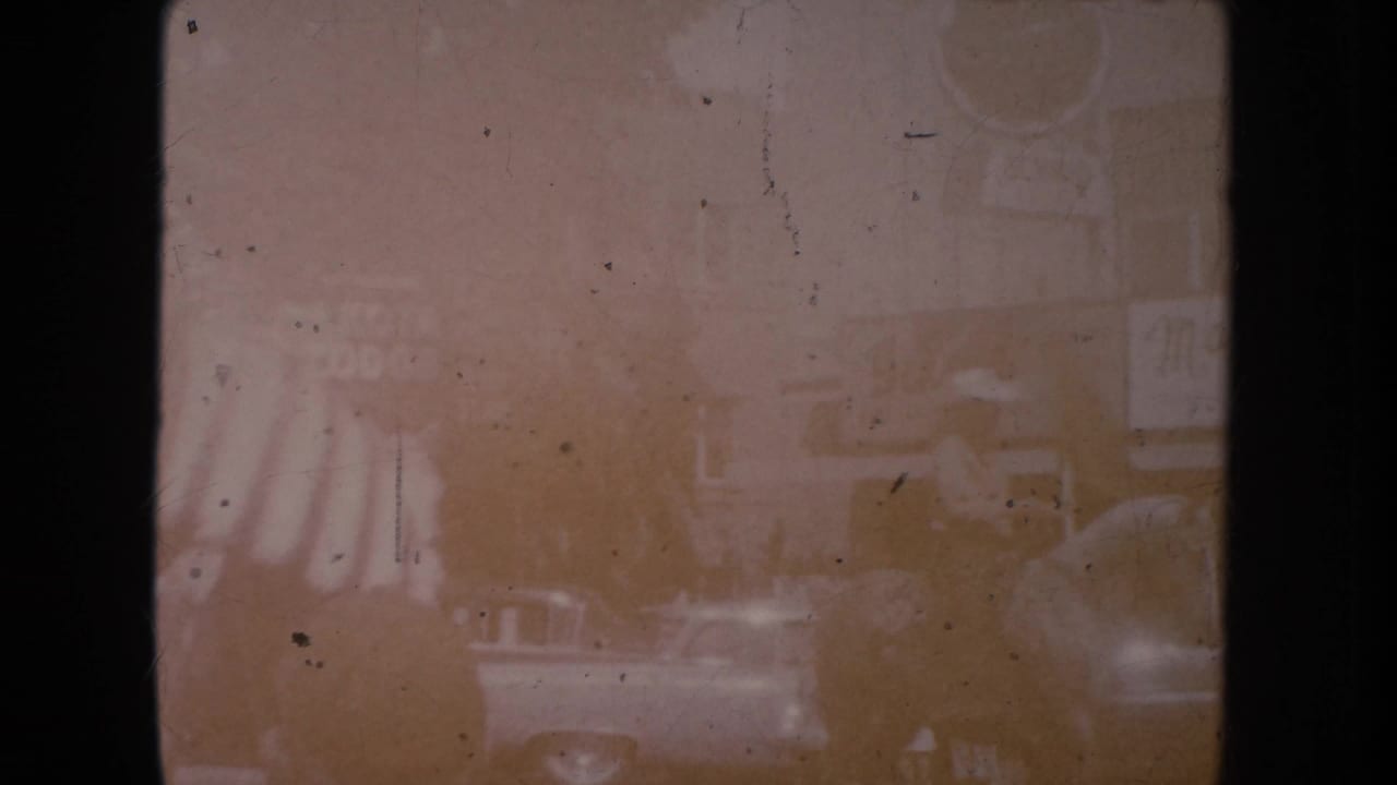 Faded street view with storefronts, parked cars, and a partial striped flag at left.