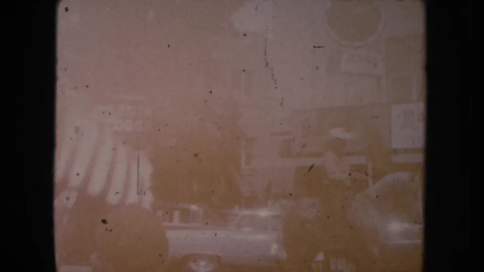 Faded 1954 South Dakota home-movie frame showing a town street with storefronts, cars, and a partial U.S. flag near the left edge.