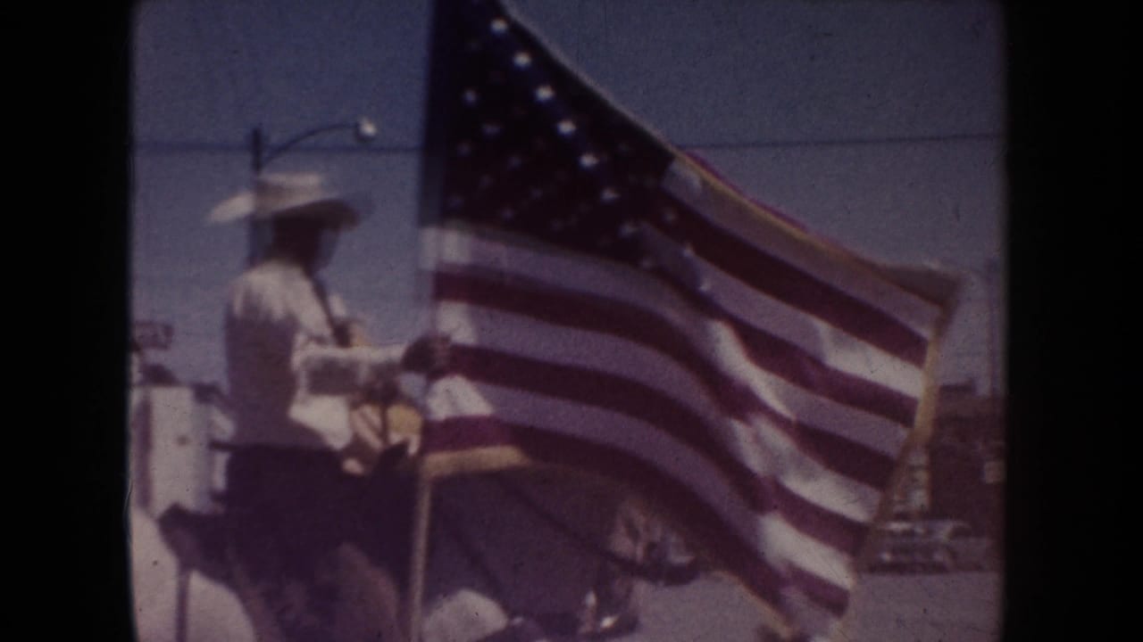 Mounted rider in a white uniform and cowboy hat carrying a large U.S. flag.