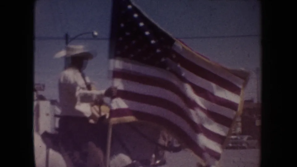 A uniformed rider in a cowboy hat passes on horseback in South Dakota, carrying a large U.S. flag in a 1954 parade scene.