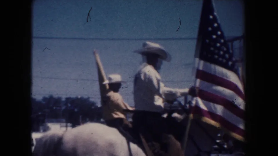Two mounted figures, including one smaller rider, move through a 1954 South Dakota parade with a large U.S. flag in the foreground.