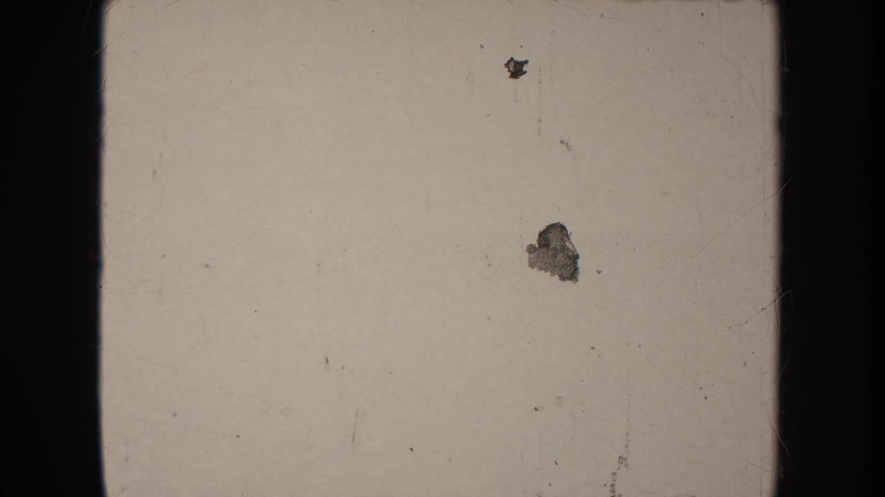 A pale 8mm frame with dark borders, visible dust, scratches, and small marks before the parade action begins.