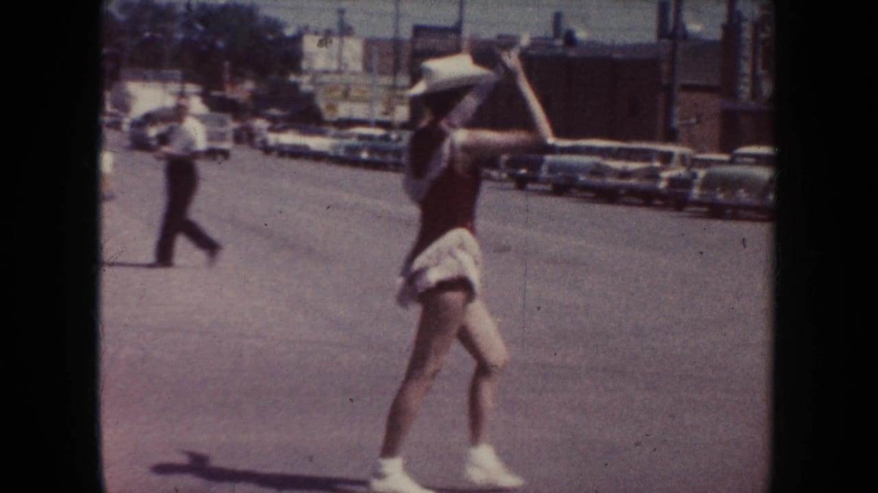 In 1954 South Dakota parade footage, a young woman in a white cowboy hat and red outfit dances in the street near parked cars.
