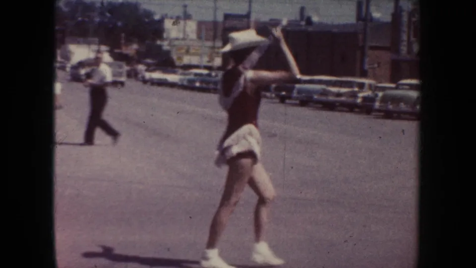 In 1954 South Dakota parade footage, a young woman in a white cowboy hat and red outfit dances in the street near parked cars.