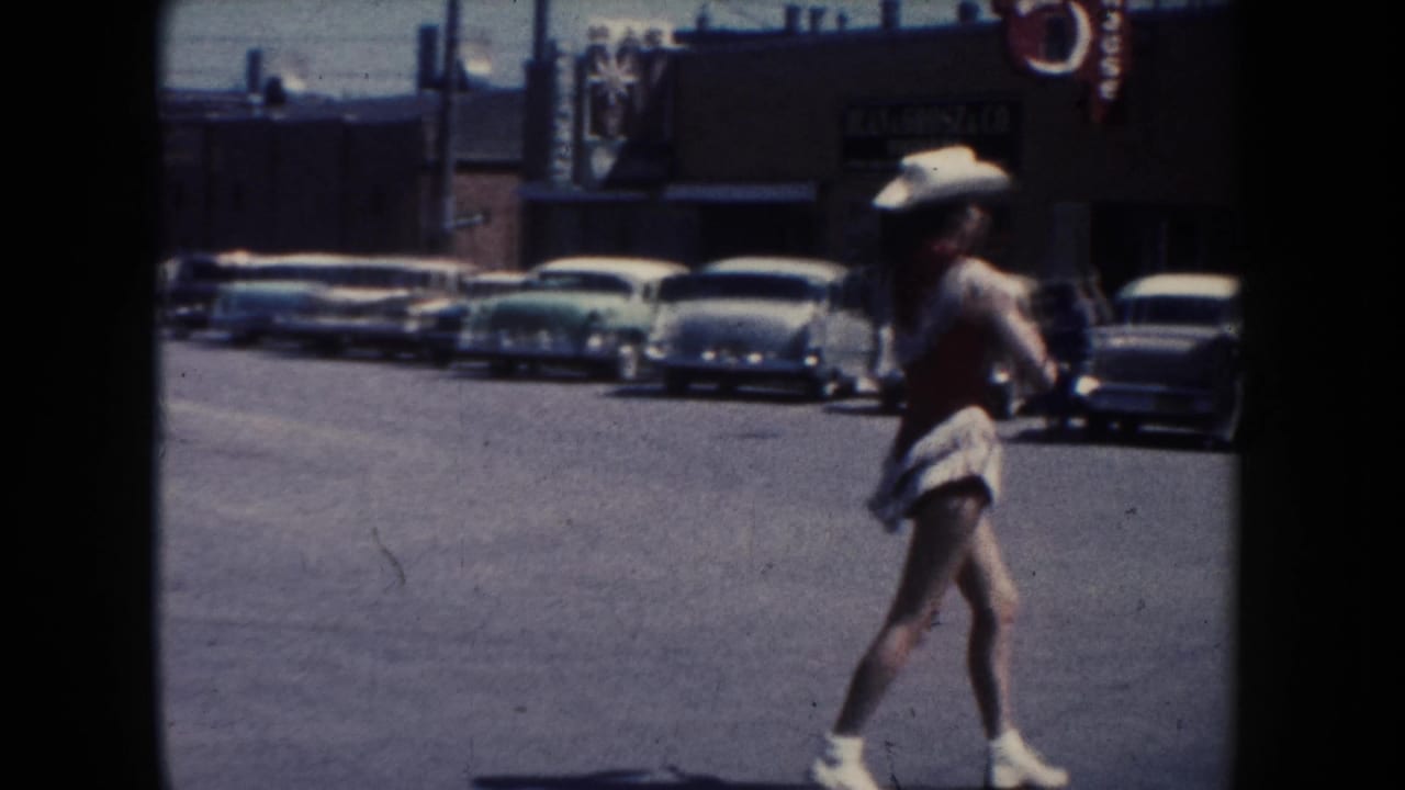 The dancer continues along a South Dakota parade street, with 1950s cars, business facades, and dark film borders in view.
