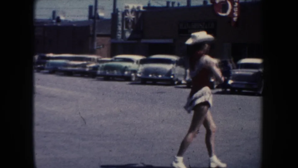The dancer continues along a South Dakota parade street, with 1950s cars, business facades, and dark film borders in view.