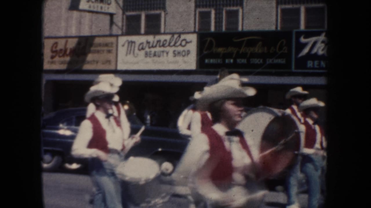 Band members in white hats and red vests march with drums past storefront signs including Marinello Beauty Shop during the 1954 parade.