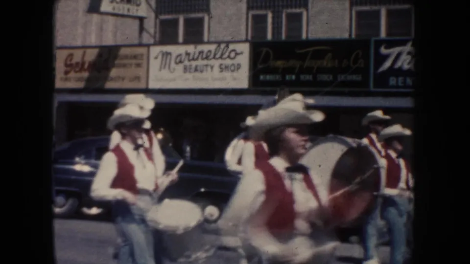 Band members in white hats and red vests march with drums past storefront signs including Marinello Beauty Shop during the 1954 parade.