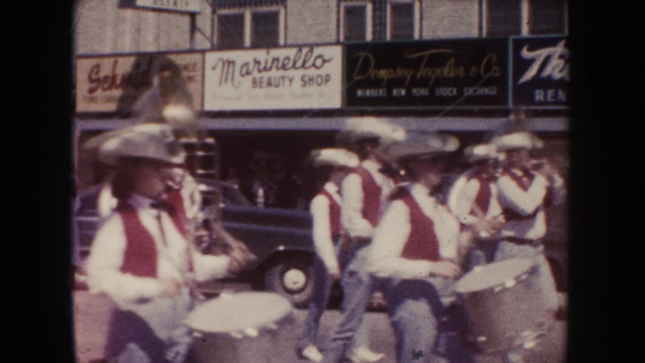 Marching band members in cowboy hats pass storefronts with brass and percussion instruments.