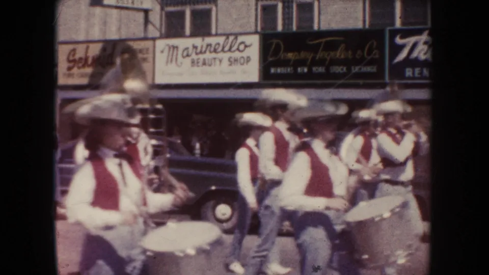 1954 South Dakota parade footage showing band members in white shirts, red vests, and cowboy hats marching past storefront signs.