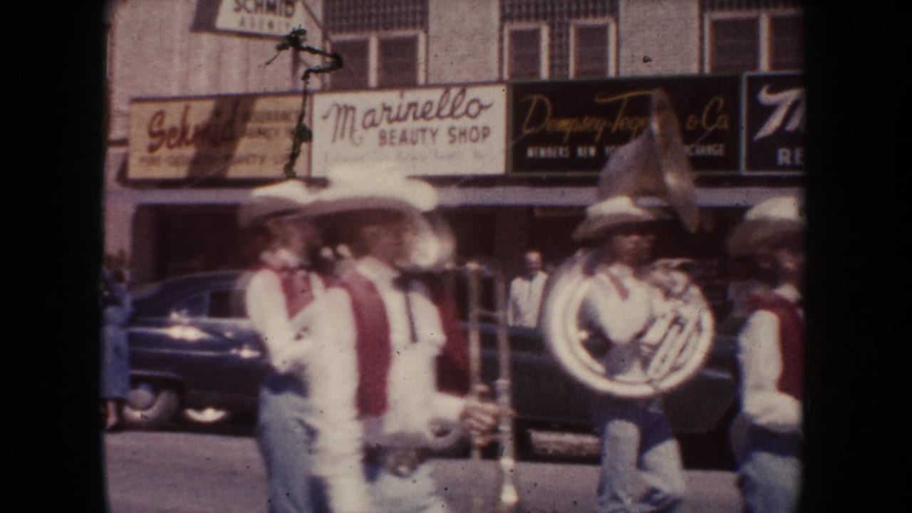 1954 South Dakota street parade view with brass musicians, a sousaphone or tuba, and mid-century cars near storefront signs.