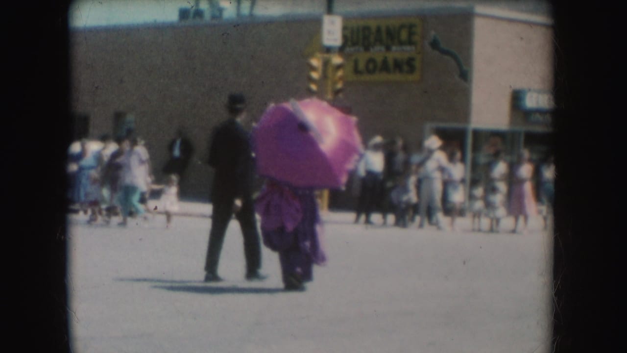 In 1954 South Dakota footage, a suited man walks with a woman holding a pink parasol near pedestrians and an Insurance Loans sign.