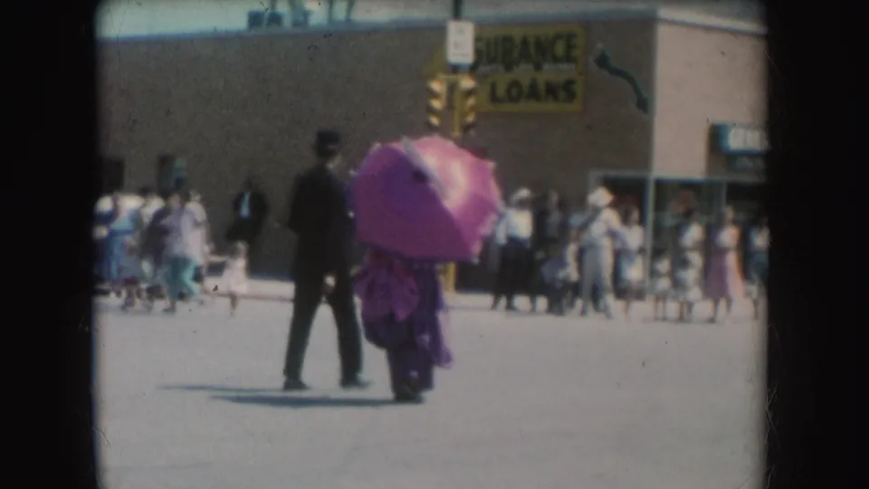 In 1954 South Dakota footage, a suited man walks with a woman holding a pink parasol near pedestrians and an Insurance Loans sign.
