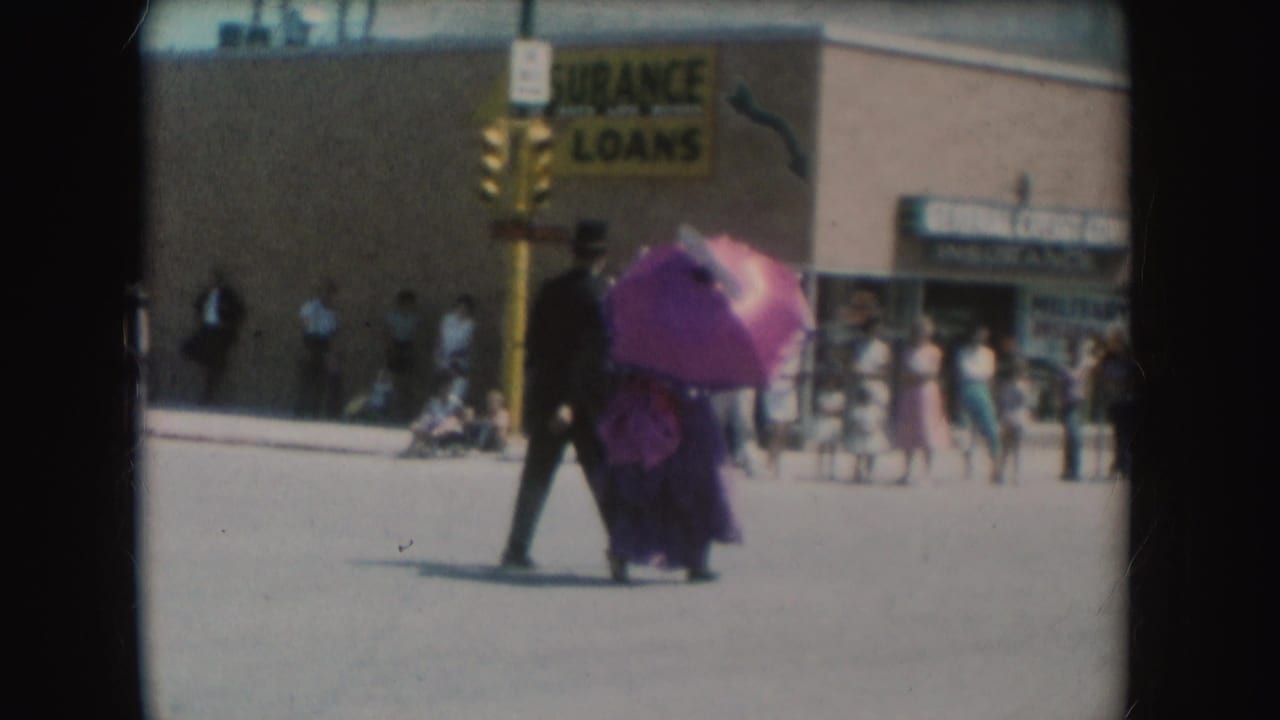 The man and woman with the pink parasol continue across a South Dakota street, with storefront signage and pedestrians in the background.