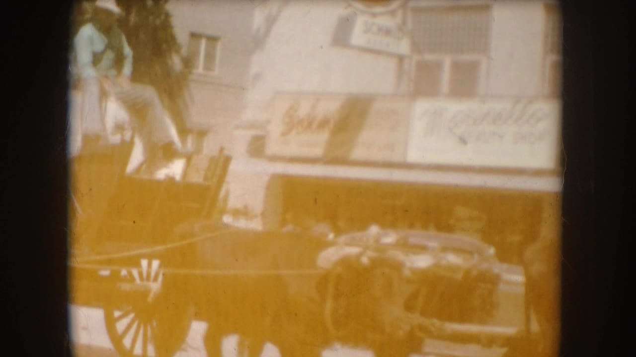 1954 South Dakota 8mm frame showing a horse-drawn carriage moving past downtown storefronts with passengers visible.