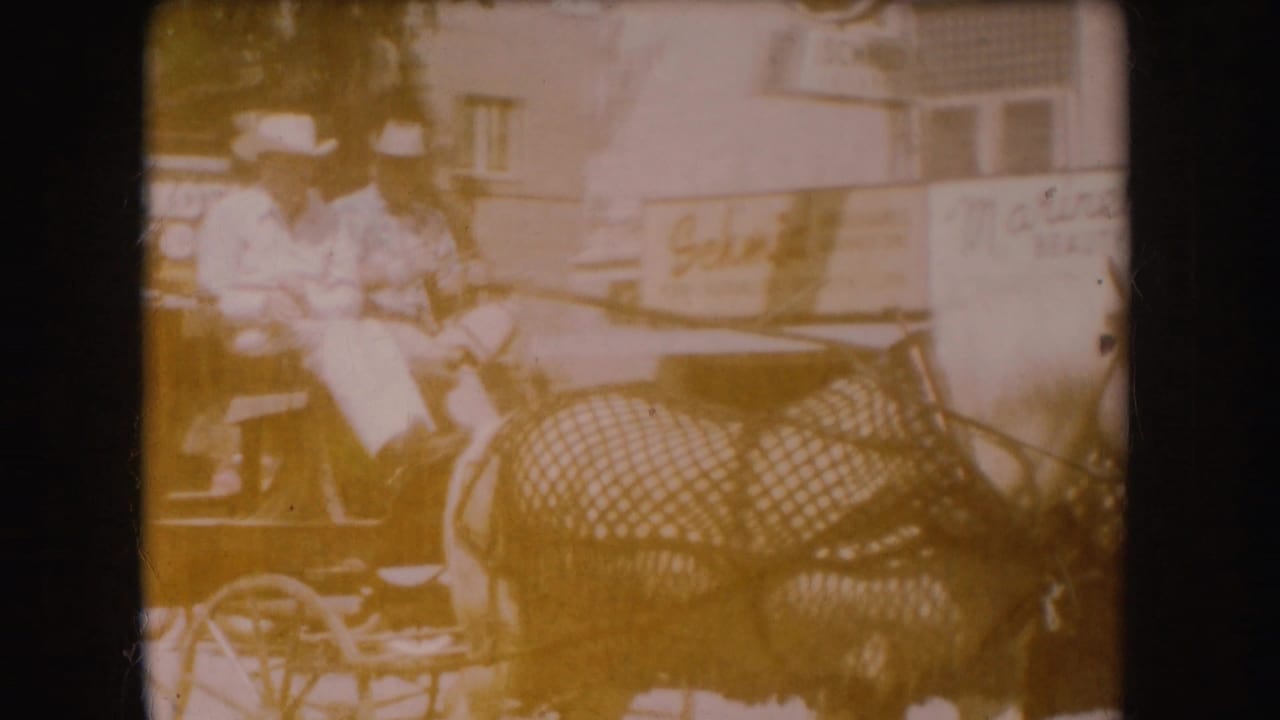 1954 South Dakota 8mm frame showing two carriage riders in hats passing storefront signs on a downtown street.