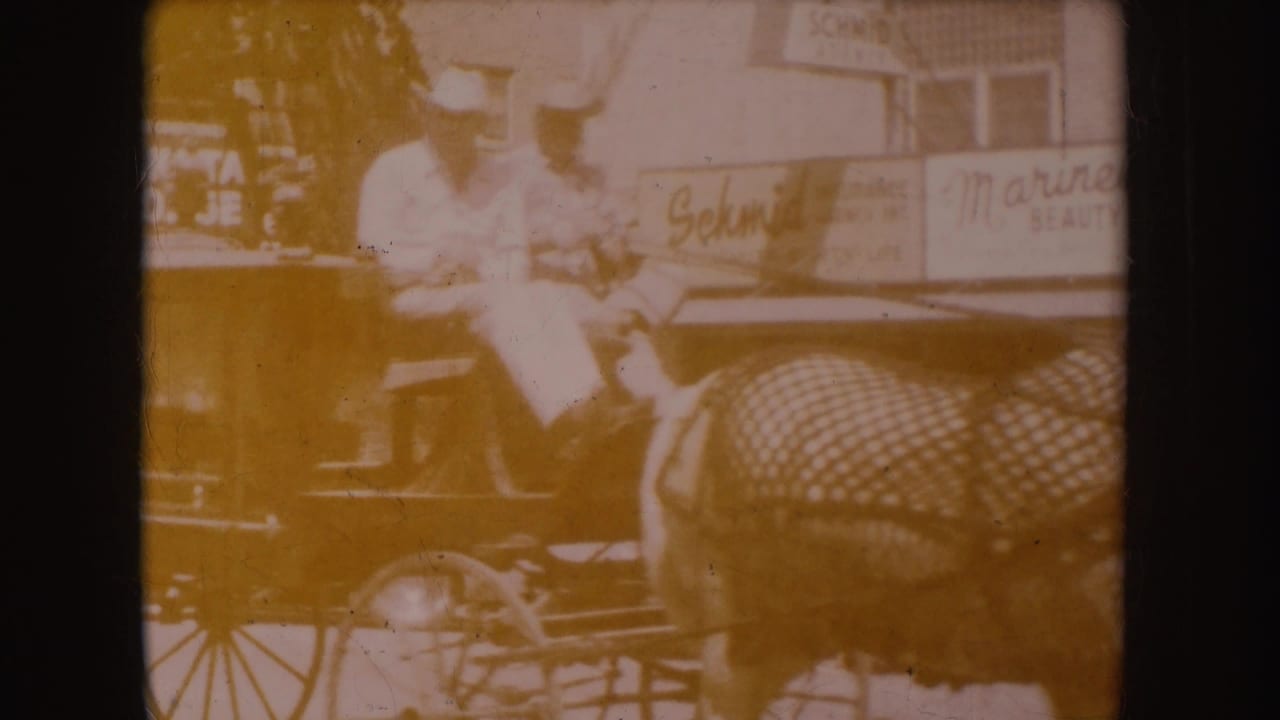 Faded frame of two men in cowboy hats seated on a horse-drawn carriage near storefronts.