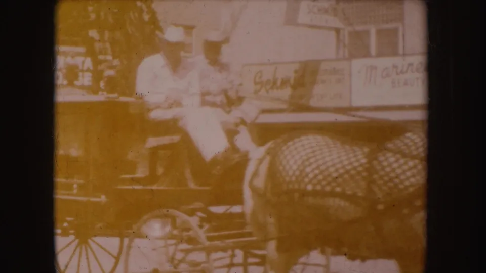 1954 South Dakota home-movie frame showing two men in cowboy hats on a horse-drawn carriage passing storefront signs.