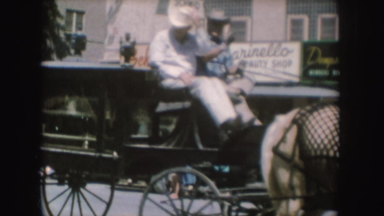 1954 South Dakota frame with two men seated on a horse-drawn carriage or hearse, with a horse and beauty shop signage behind them.