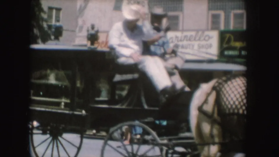 1954 South Dakota frame with two men seated on a horse-drawn carriage or hearse, with a horse and beauty shop signage behind them.