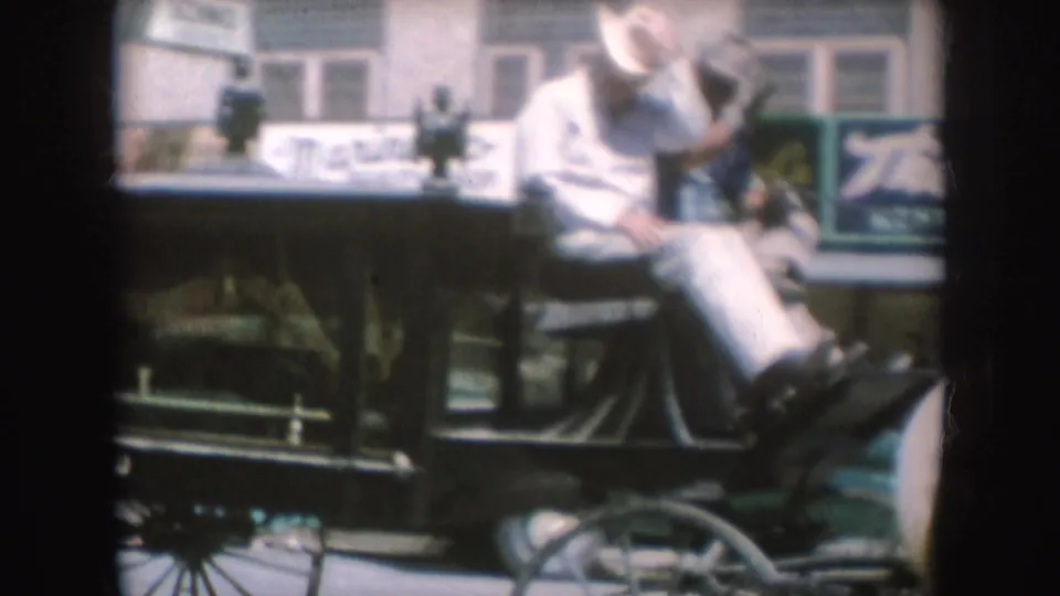 1954 South Dakota home-movie frame showing a cowboy-hatted rider on the front bench of a horse-drawn carriage in town.