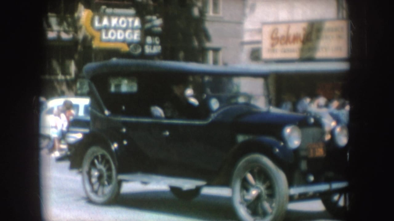 Black vintage convertible passing storefronts with lodge and insurance signs.