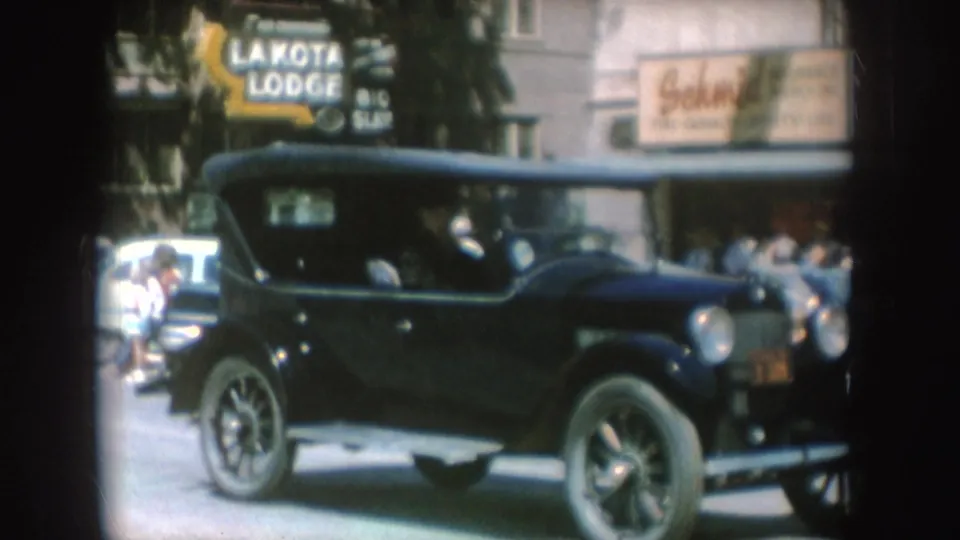 1954 South Dakota 8mm frame showing a black vintage convertible on a town street beside visible storefront signs.