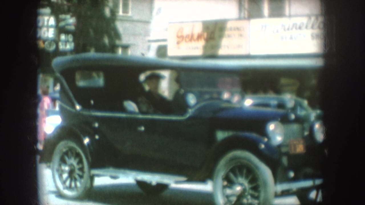 1954 South Dakota home-movie frame with a dark vintage convertible passing commercial storefronts and partially readable signs.