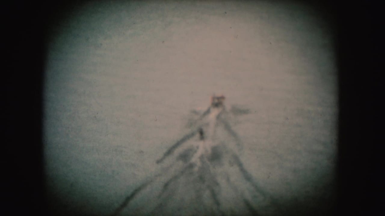 1950s home-movie frame of a distant boat and water-skier shape on a river, softened by grain and exposure falloff.