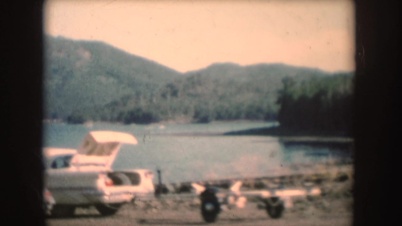 White 1950s car and small trailer parked beside a lake with wooded mountains beyond.