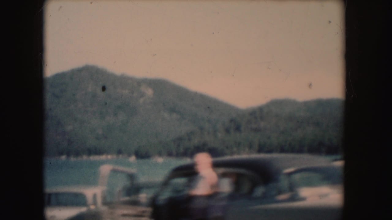 1954 South Dakota lakeside frame with a person near a dark parked car, open door, lake, and wooded hills.