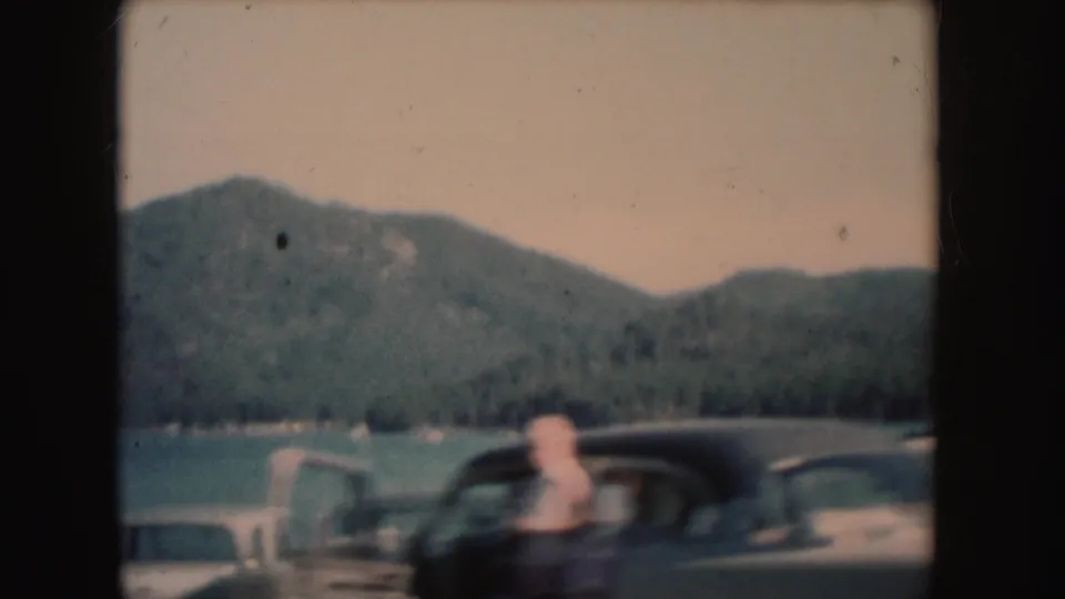 1954 South Dakota lakeside frame with a person near a dark parked car, open door, lake, and wooded hills.