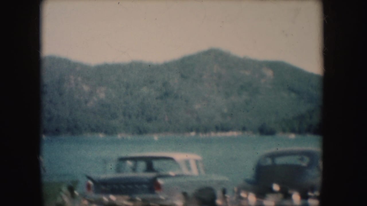 1954 South Dakota lakeside view with parked mid-century cars, blue-green water, and forested mountains.