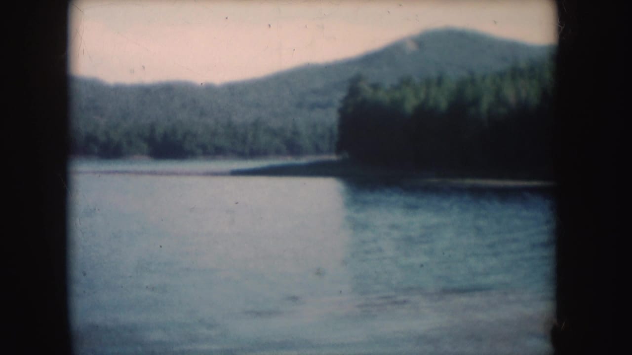 1954 South Dakota 8mm frame of a quiet lake bordered by forest and hills, with a small distant boat on the water.