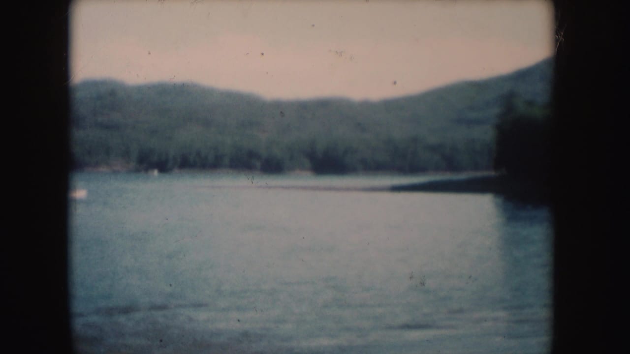 1954 South Dakota 8mm frame showing a broad lake surface with forest along the far shore and low hills behind it.