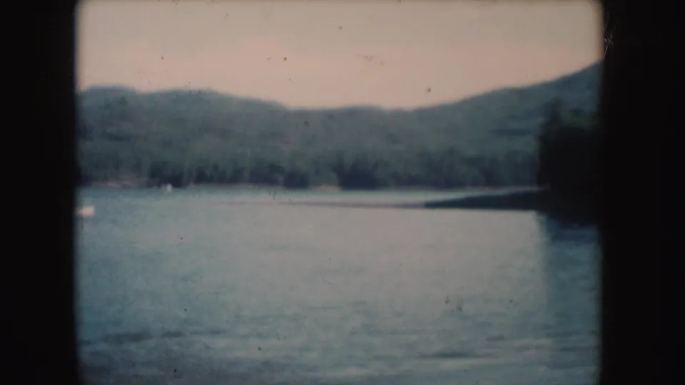 1954 South Dakota 8mm frame showing a broad lake surface with forest along the far shore and low hills behind it.