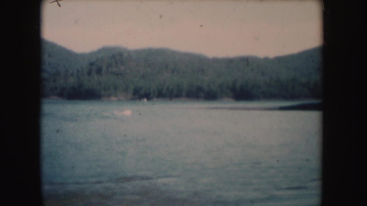 1954 South Dakota 8mm frame with a small distant boat on calm water, backed by forested shoreline and rolling hills.