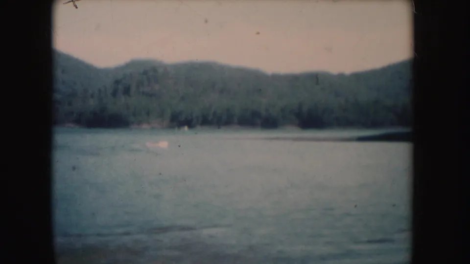 1954 South Dakota 8mm frame with a small distant boat on calm water, backed by forested shoreline and rolling hills.