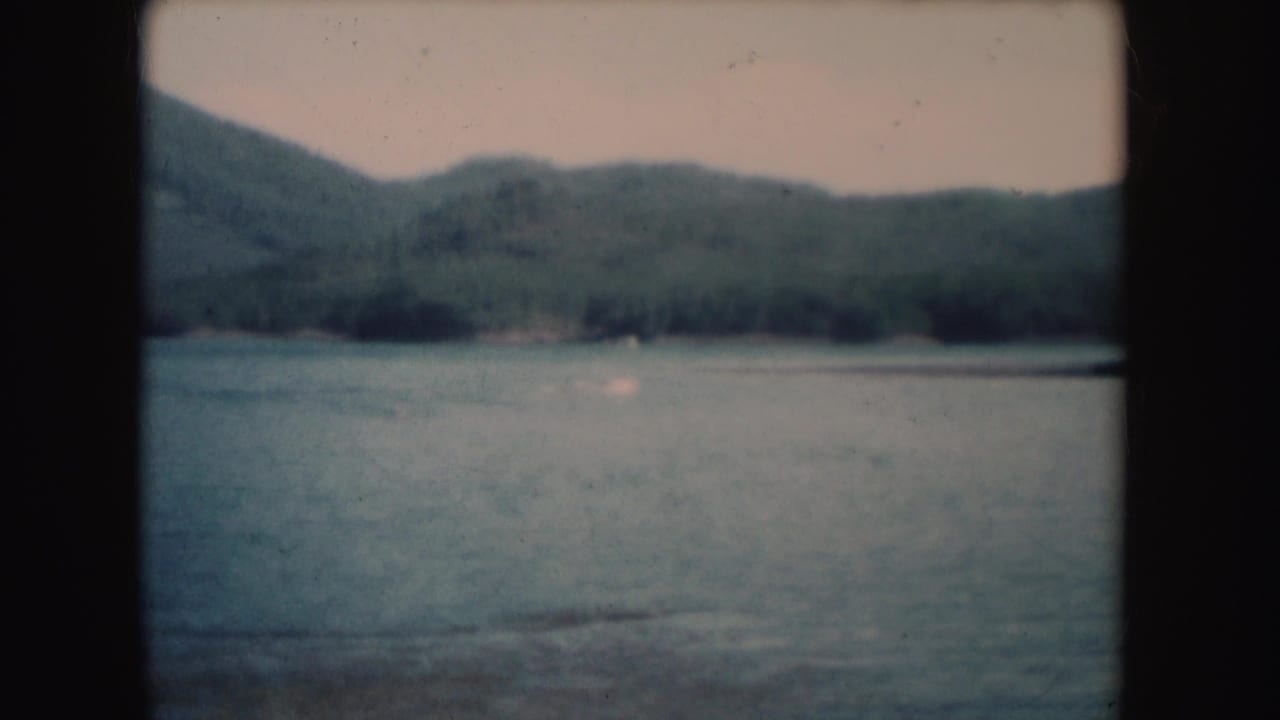1954 South Dakota 8mm frame showing a calm lake, distant forested hills, soft sky, and visible film gate borders.