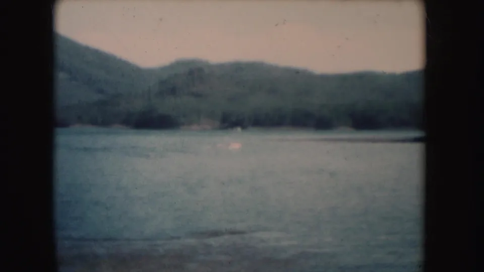 1954 South Dakota 8mm frame showing a calm lake, distant forested hills, soft sky, and visible film gate borders.