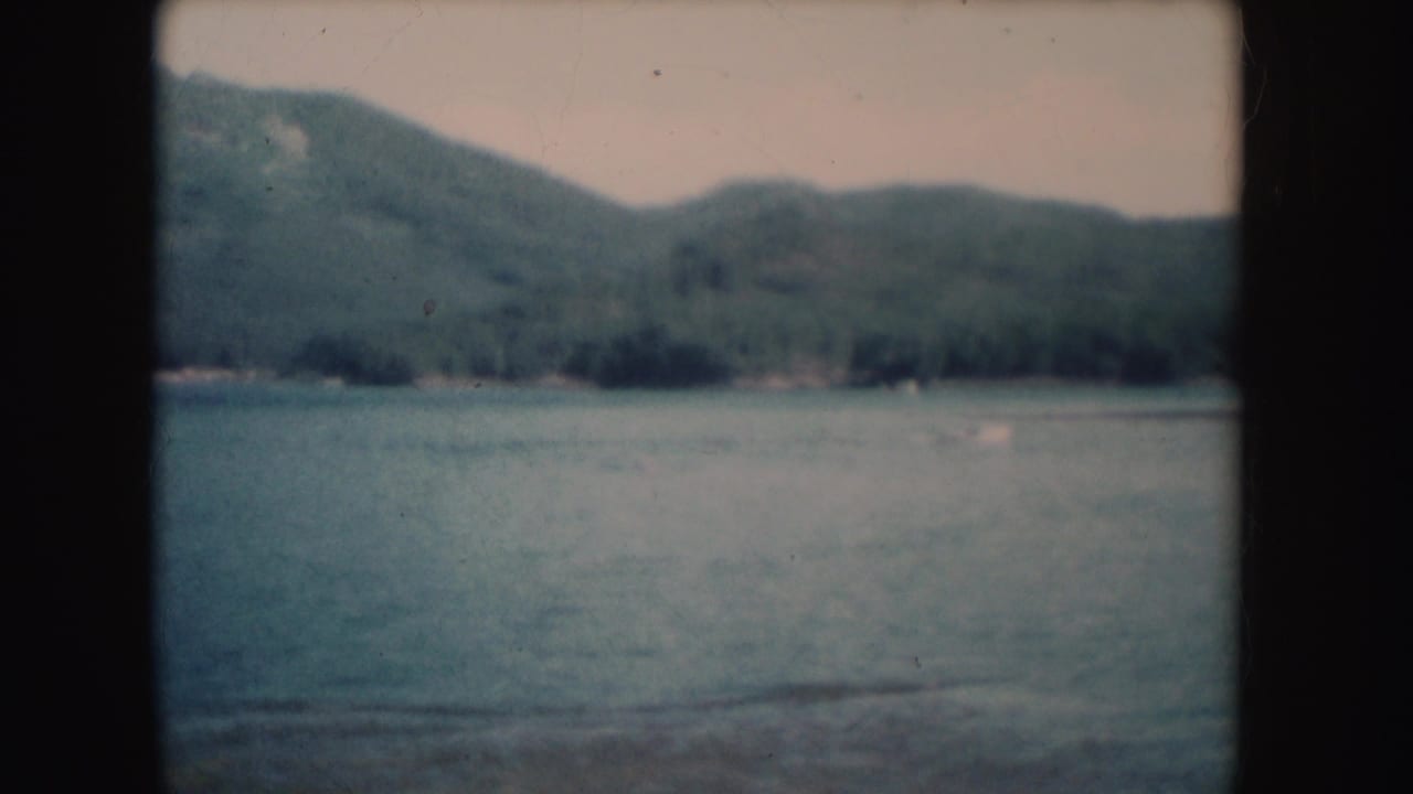 1954 South Dakota home-movie frame of lake water below wooded hills, with a faint distant shoreline and dark film borders.