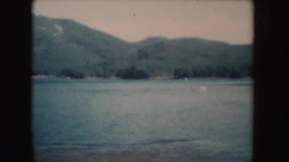 1954 South Dakota home-movie frame of lake water below wooded hills, with a faint distant shoreline and dark film borders.