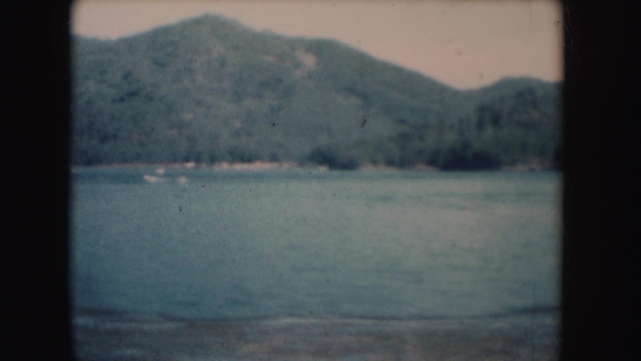 1954 South Dakota 8mm frame showing a lake, wooded hillside, and a small pale boat near the far shoreline.