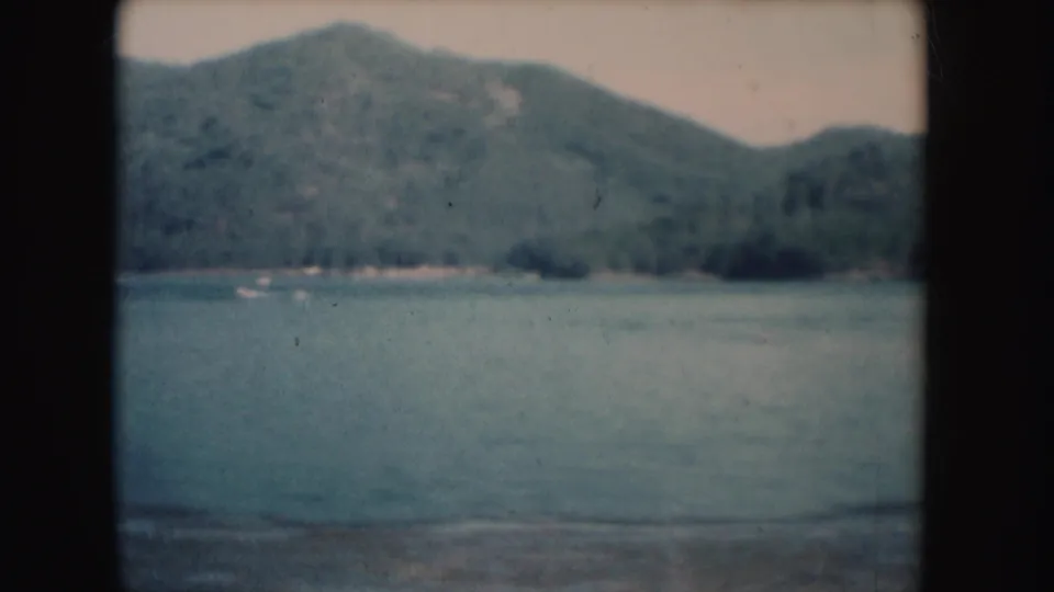 1954 South Dakota 8mm frame showing a lake, wooded hillside, and a small pale boat near the far shoreline.