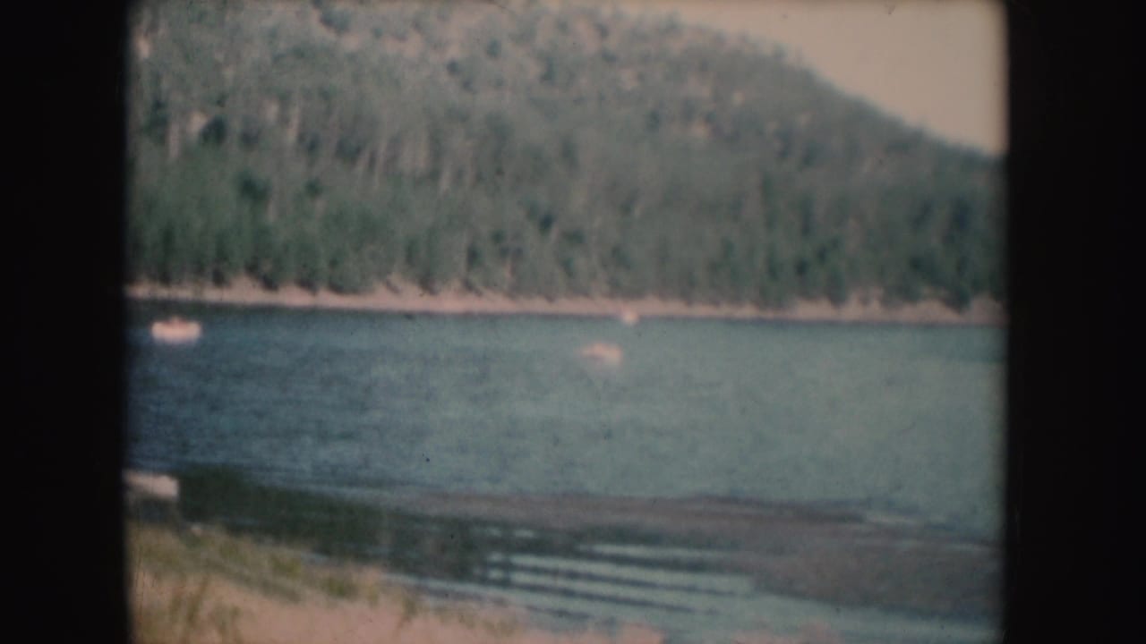 1954 South Dakota frame showing rippled lake water, a forested slope, shoreline, and small distant boats near the far bank.