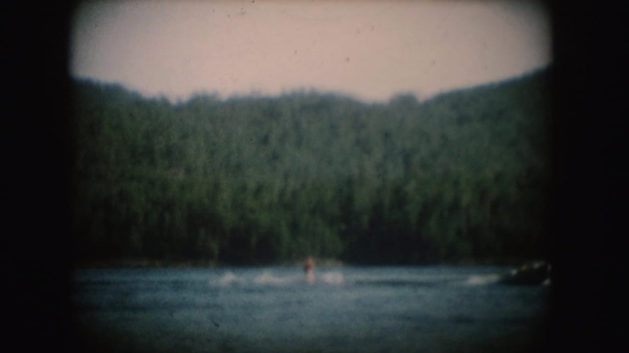 Distant water skier on a South Dakota lake, pulled near a motorboat against wooded hills in 1954 home-movie footage.