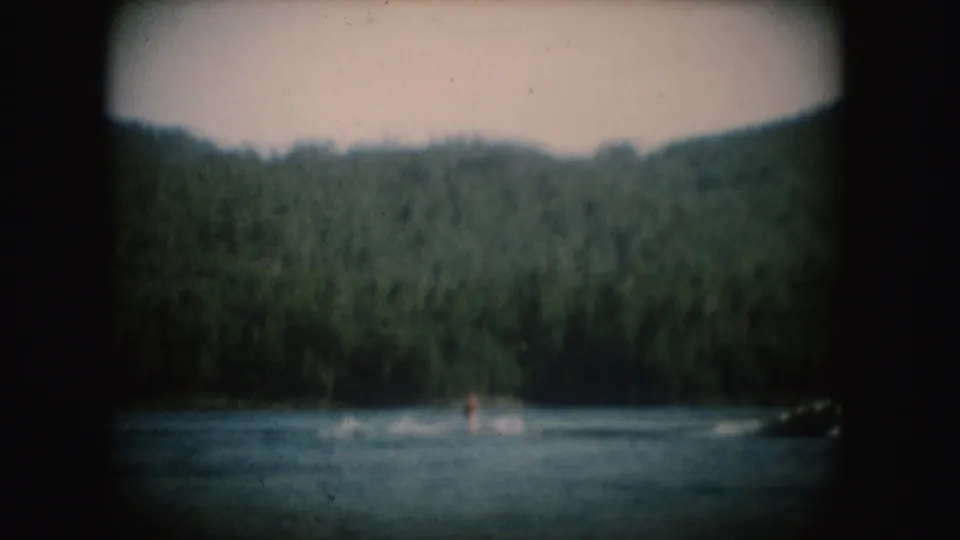 Distant water skier on a South Dakota lake, pulled near a motorboat against wooded hills in 1954 home-movie footage.